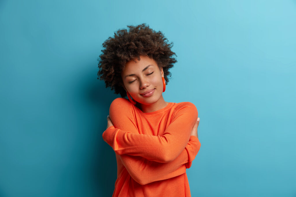 Self care and tenderness concept. Pleased dark skinned woman embraces herself, holds shoulders, feels comfort in her new sweater, has romantic tender mood, needs feel warmth, comfort and love