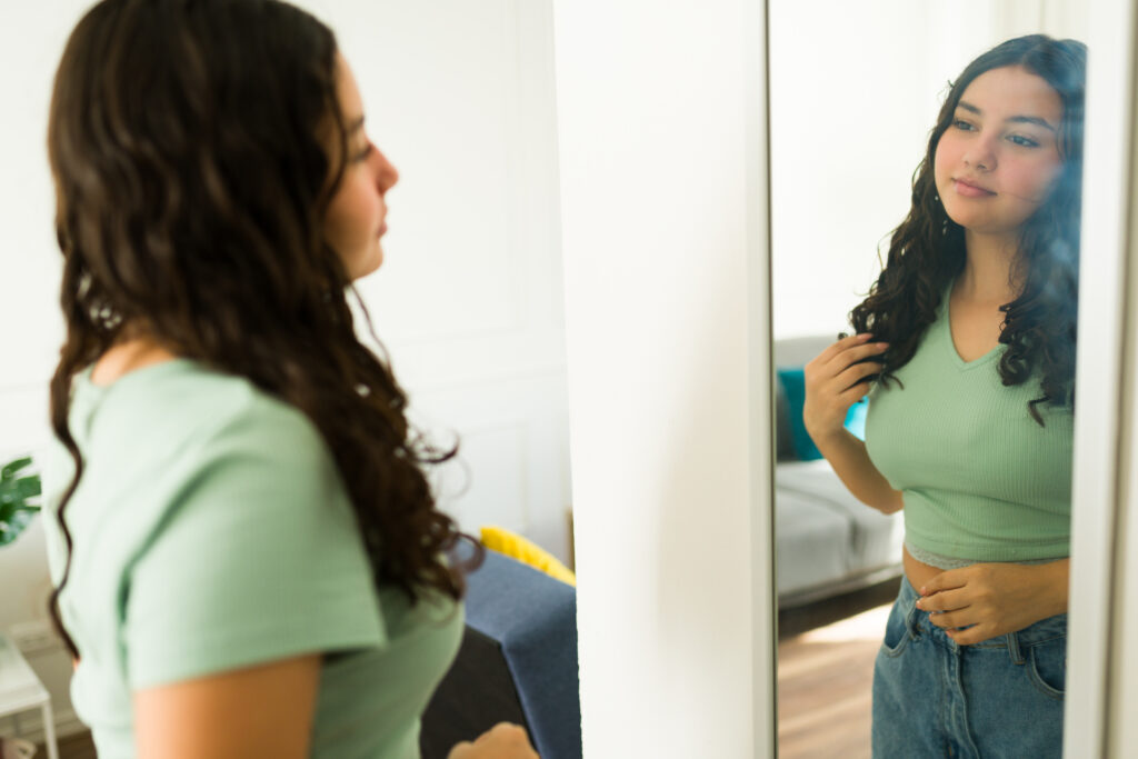 Beautiful latin teenager girl admiring herself in a mirror at home, touching her hair and smiling